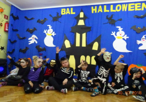 Na tle dekoracji halloweenowej siedzą dzieci. Niektóre dzieci podnoszą rękę w górę.